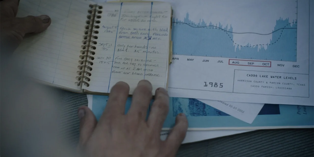 Caddo Lake (2024) movie still: closeup — A cluttered desk surface holds a spiral-bound notebook and a printed water level chart. A human hand rests…