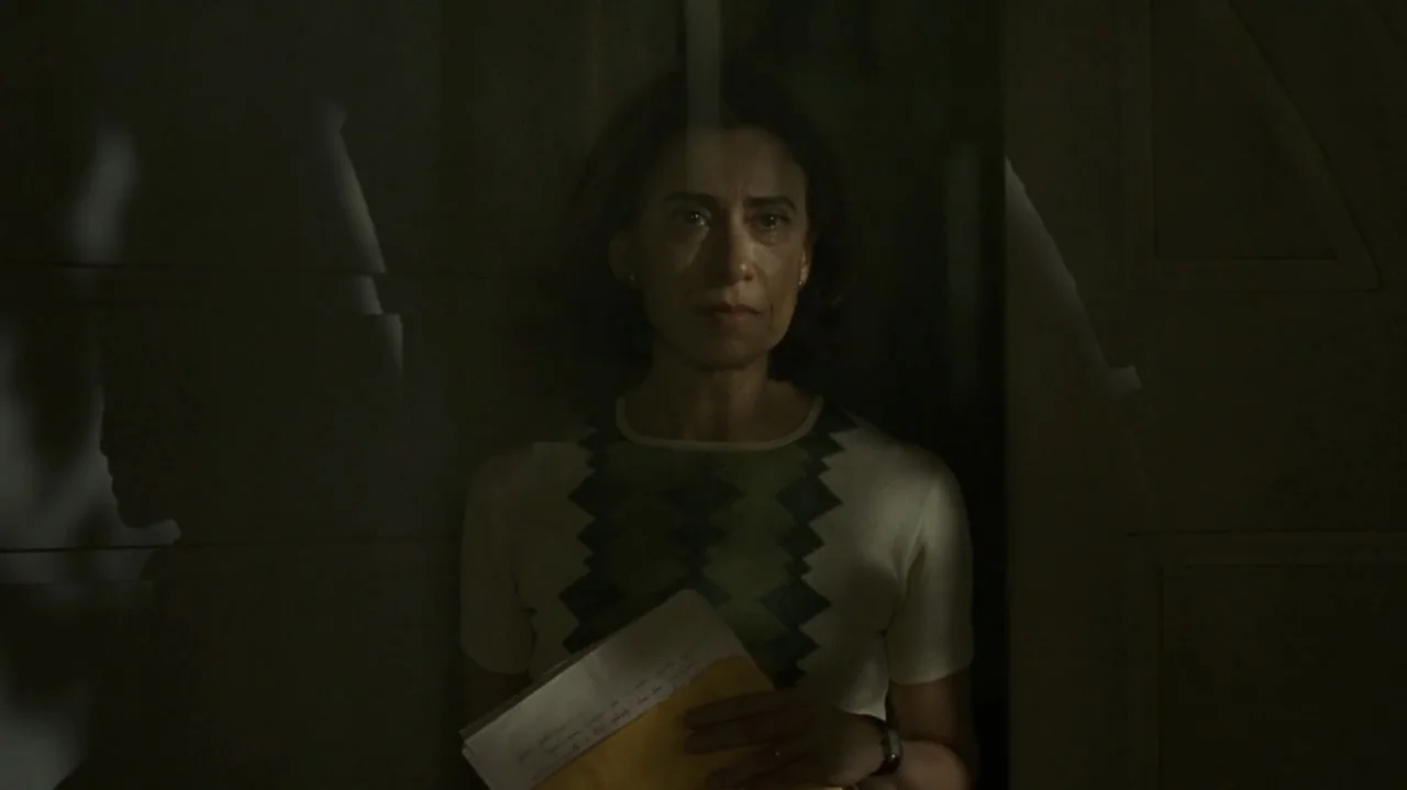 I'm Still Here (2024) movie still: medium — A dimly lit indoor hallway with dark paneled walls and reflective glass surfaces. A woman stands holding…