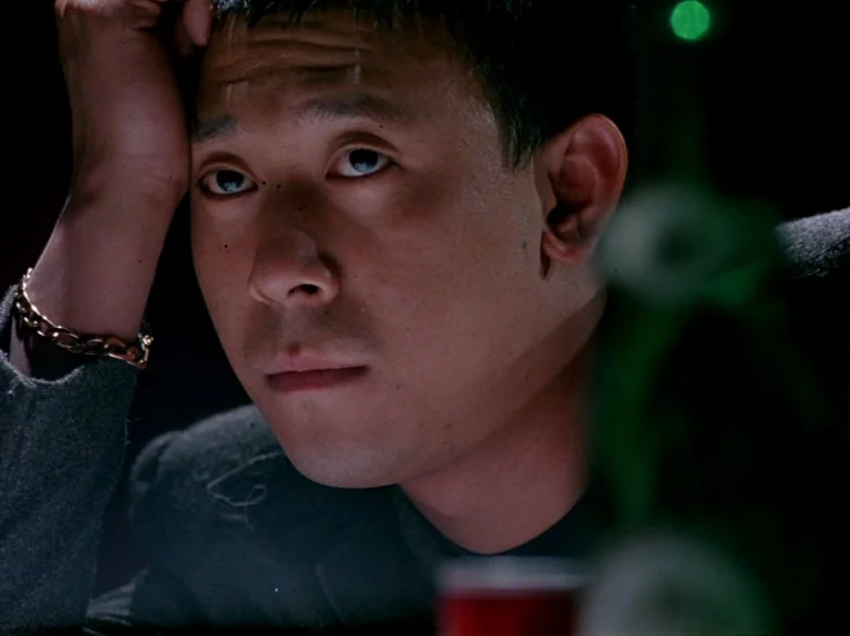 Black Snow (1990) movie still: closeup — A dimly lit space features a man leaning his head against his hand. He sits in the foreground with a…