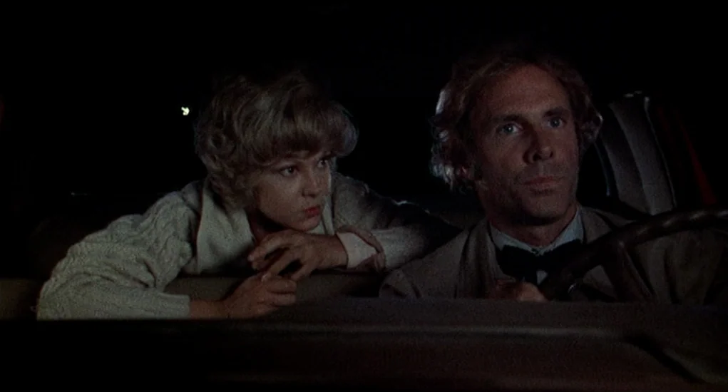 Family Plot (1976) movie still: medium — The interior of a car at night with two occupants sitting in the front seats. A man is behind the wheel,…