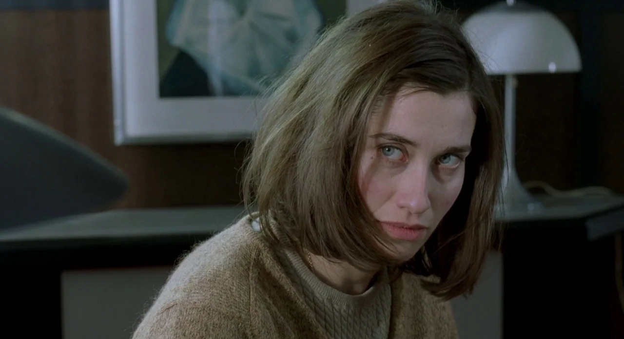 Read My Lips (2001) movie still: closeup — A woman stands in a professional interior space with wood paneling and office furniture. The subject…