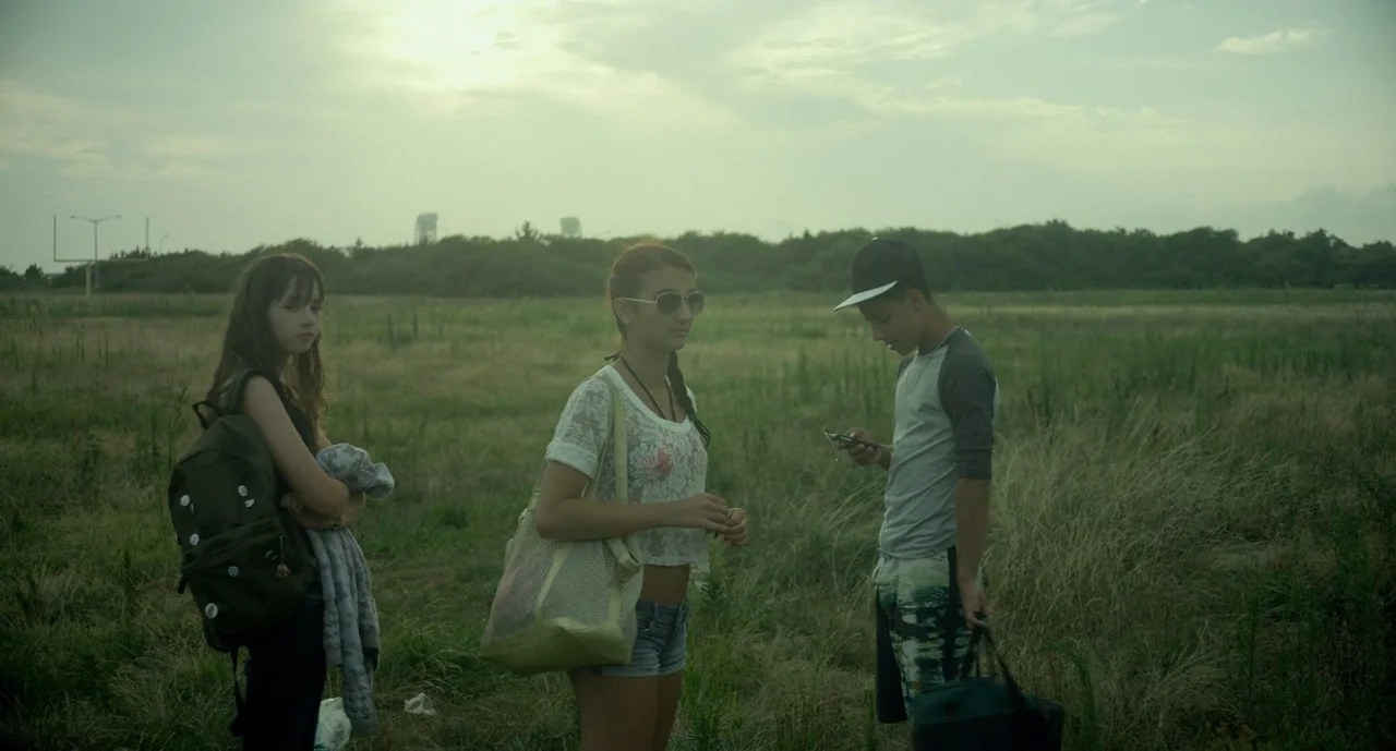 It Felt Like Love (2013) movie still: fullbody — An overcast open field with tall grass stretches toward a line of distant trees. Three teenagers…