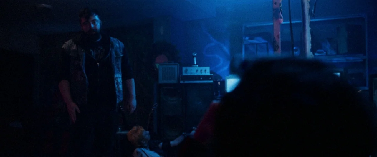 VFW movie still: medium — A dark, blue-lit basement filled with electronic equipment and speakers. A man stands over a woman sitting low in the…