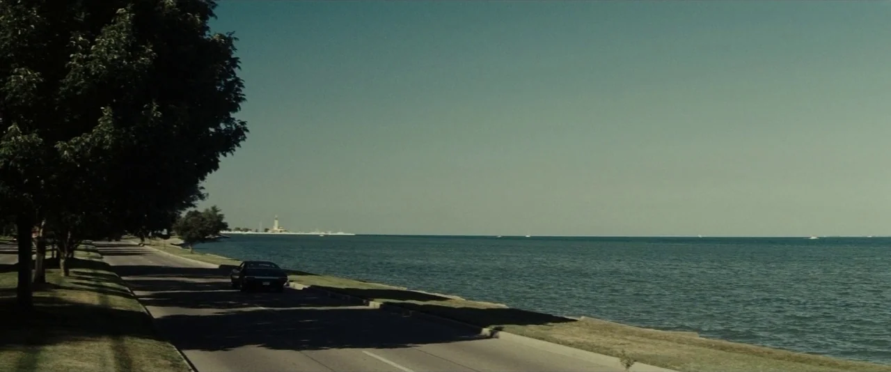 Gran Torino (2008) movie still: wide — A dark car travels along an asphalt road bordered by trees and a large lake. A distant structure is visible…