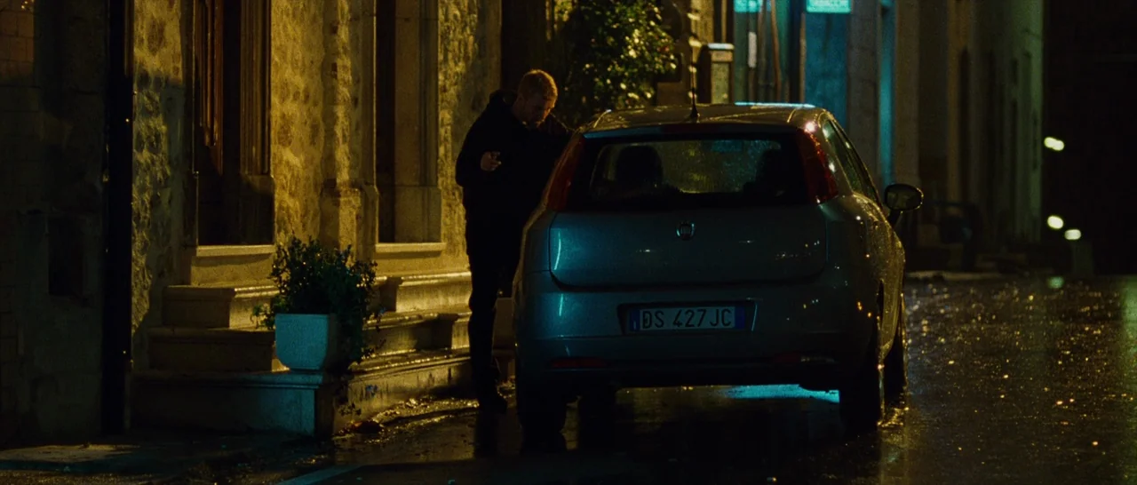 The American (2010) movie still: fullbody — A wet asphalt road at night lined by stone building facades and a parked car. The man stands near the…