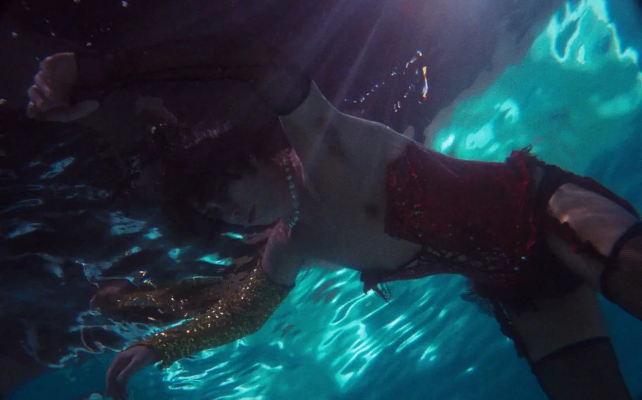 The Rocky Horror Picture Show (1975) movie still: medium — A person floats underwater in a pool while wearing a red corset and gold sleeves.…