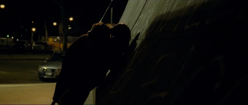 Shame (2011) movie still: medium — A dimly lit parking lot at night features a large, graffiti-covered concrete wall. Two people stand in shadow…