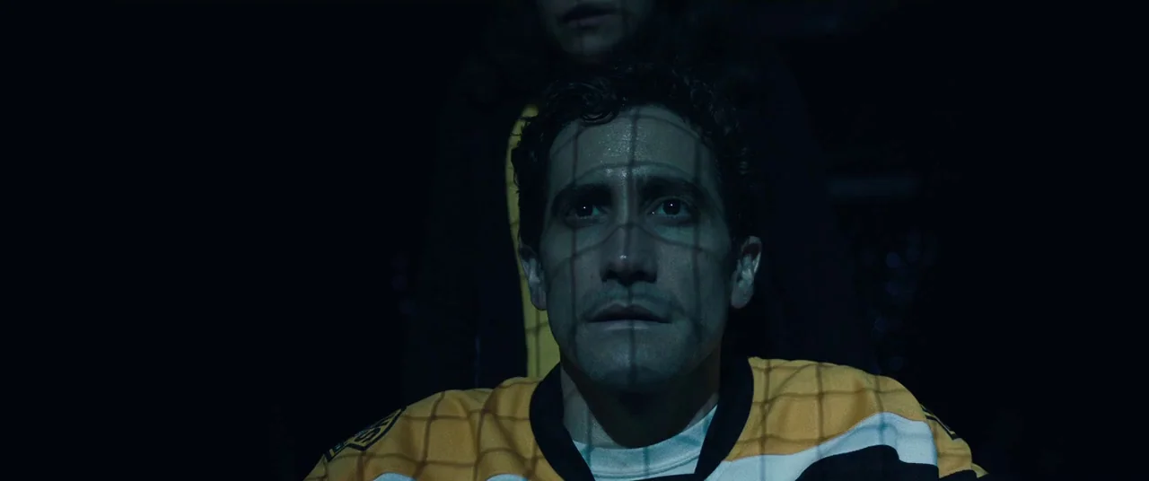 Stronger (2017) movie still: closeup — A man wearing an athletic jersey sits in a dark, shadow-filled environment. A grid pattern of light is…