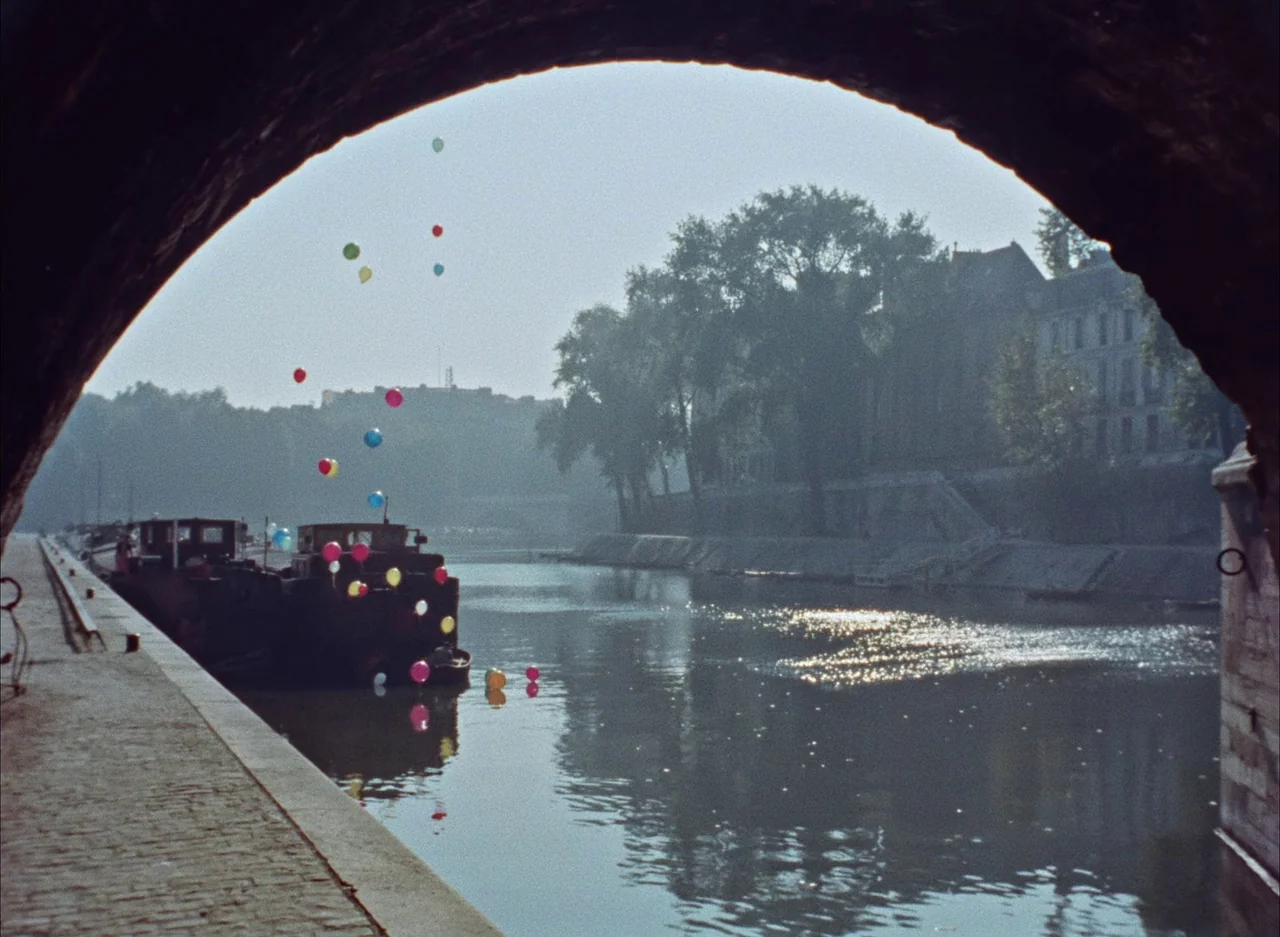 The Red Balloon (1956) movie still: establishing — A stone embankment overlooks a calm river with a docked barge and urban architecture. Many…