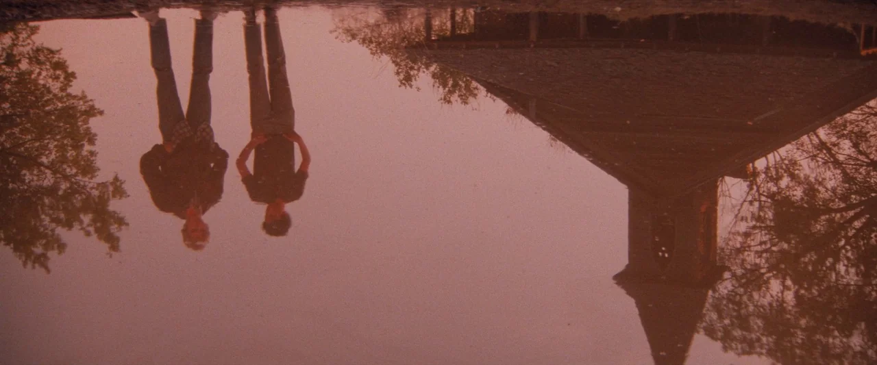 The Outsiders (1983) movie still: wide — A puddle surface displays a reflection of two people and a church building. The figures stand side by side…