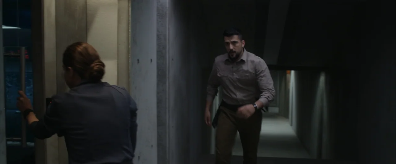 Black Site (2022) movie still: medium — A dimly lit concrete passageway features textured gray walls and a sleek modern door. A man approaches while…