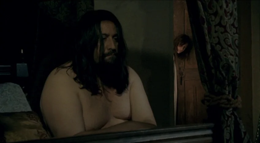 Bluebeard (2009) movie still: medium — A dimly lit room features heavy velvet curtains and wood paneling in the background. A shirtless man sits in…