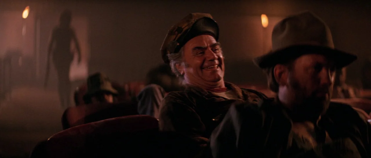 Escape From New York (1981) movie still: medium — A dimly lit room with several people seated in plush, dark red chairs. An elderly man with a cap…
