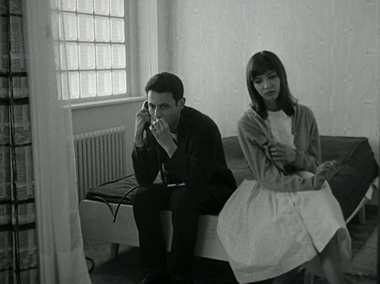 Le Petit Soldat (1963) movie still: medium — A small room features a white single bed positioned beneath a window with glass panes. A man sits on…