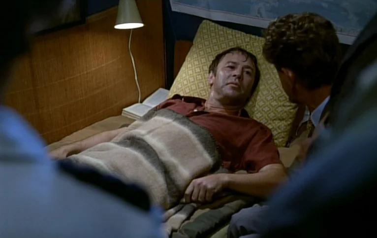 Giliap (1975) movie still: medium — A small room features a bed with a geometric yellow pillow and tan blanket. A man lies on the bed while another…