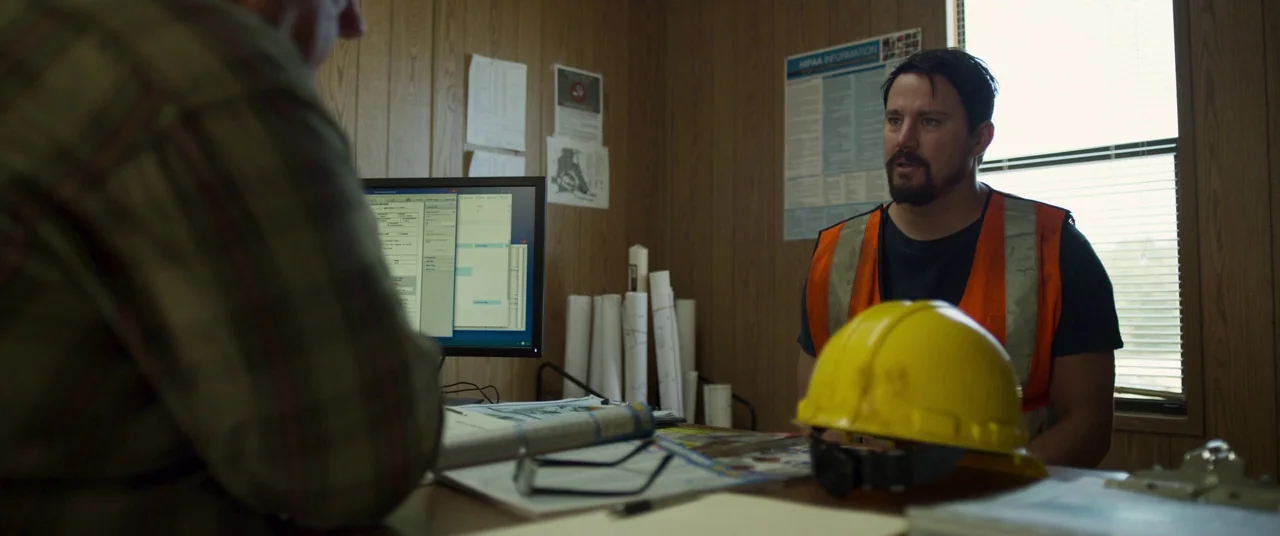 Logan Lucky (2017) movie still: medium — A small office with wood-paneled walls features a desk cluttered with blueprints and documents. A man in a…