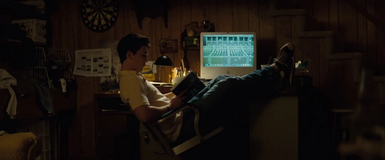 The Spectacular Now (2013) movie still: medium — A dark basement features wood-paneled walls and a cluttered desk area with a dartboard. A young man…