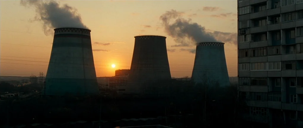 Elena (2011) movie still: wide — Three large industrial cooling towers stand in a row under a setting sun. A multi-story apartment block occupies…