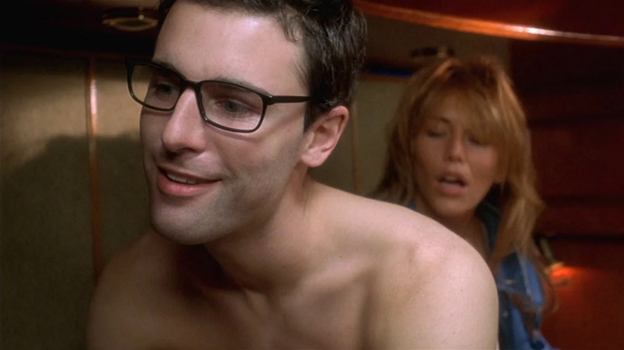 Dagon (2001) movie still: medium — A dimly lit cabin interior with wood-paneled walls and low-hanging ceiling accents. A shirtless man with glasses…
