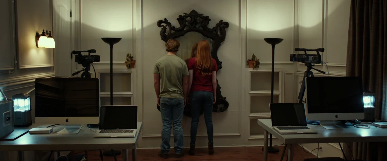 Oculus (2013) movie still: wide — A room with white wainscoting features two identical desk setups facing each other. Two people stand in the center…