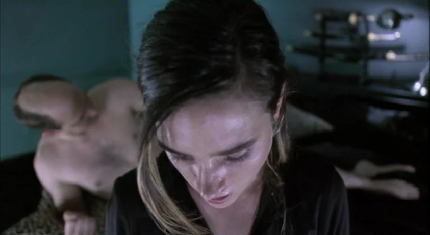 Requiem For A Dream (2000) movie still: closeup — A dark, dimly lit room contains a bed with a person sitting in front. An out-of-focus man lies on…