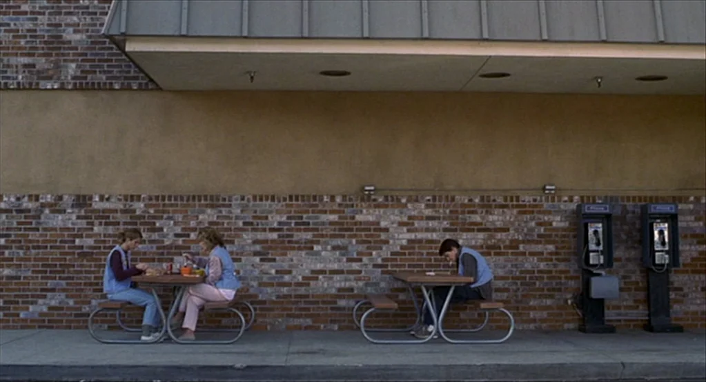 The Good Girl (2002) movie still: wide — A brick storefront facade features a recessed sidewalk area with metal picnic tables. Three people sit at…