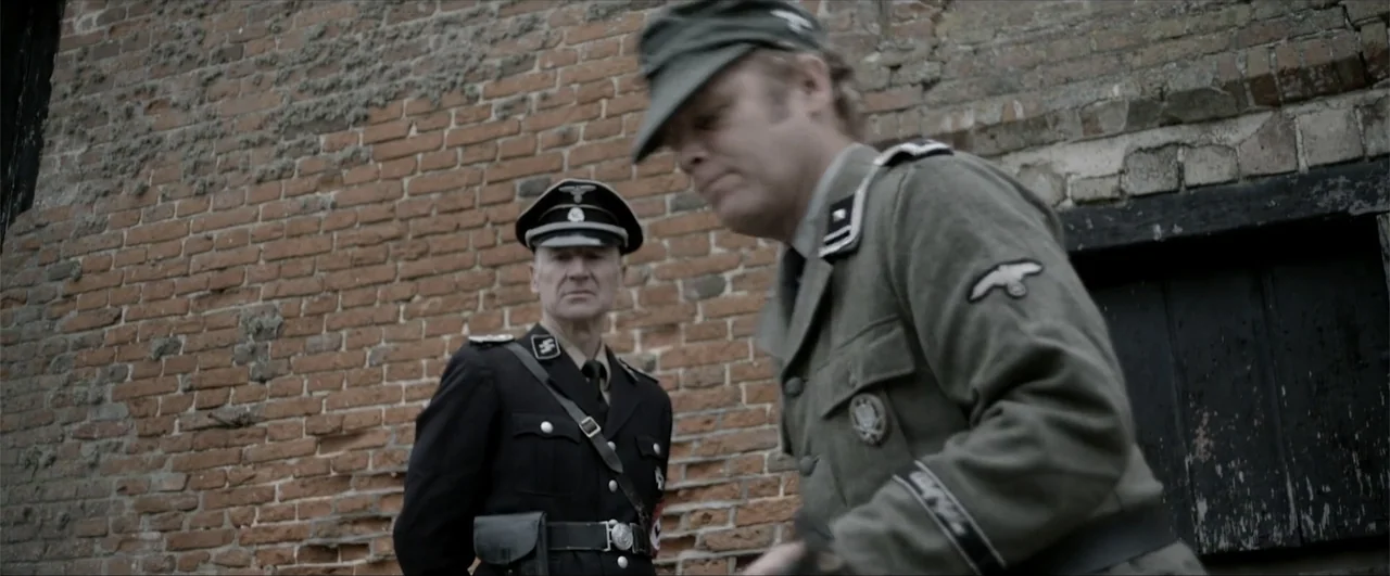 Behind the Line: Escape to Dunkirk (2020) movie still: medium — Two men in mid-century military uniforms stand against a textured red brick wall…