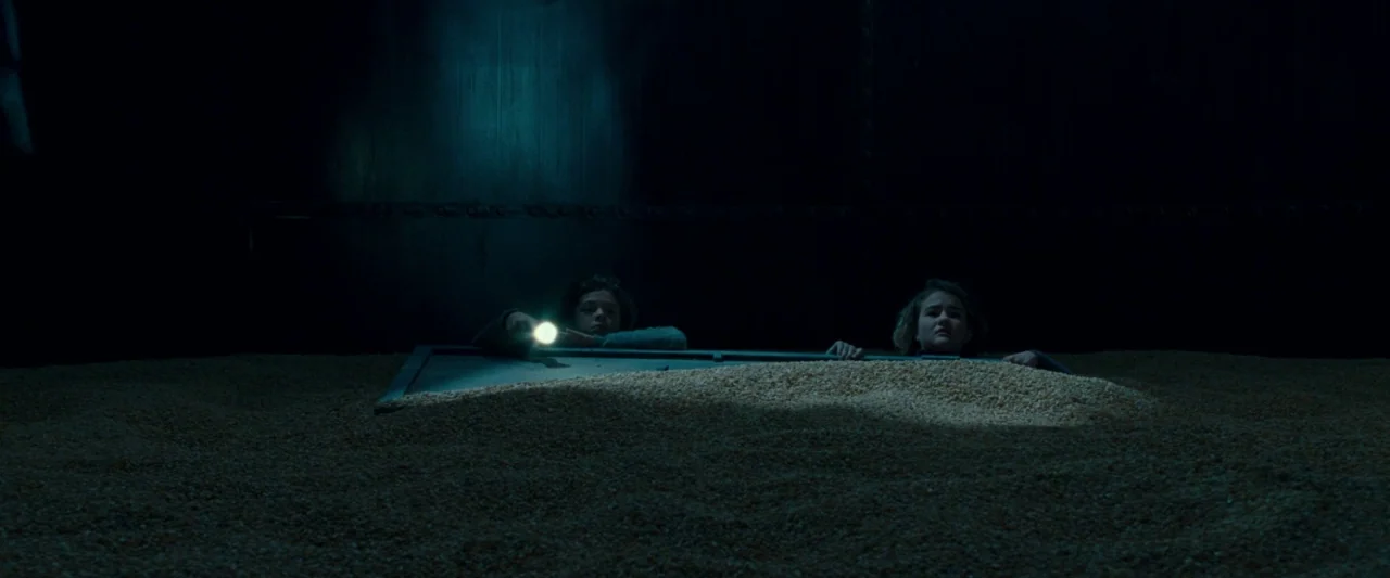 A Quiet Place (2018) movie still: wide — Two children peer over the edge of a dark, metallic grain silo filled with corn. They hold a small…