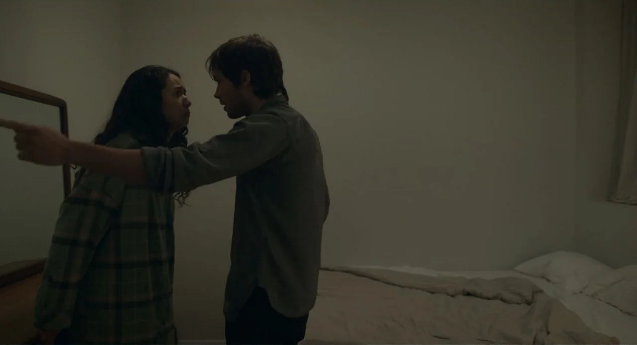 This Closeness (2024) movie still: medium — A sparsely furnished bedroom with beige walls and a bed with unmade linens. A young man and woman face…