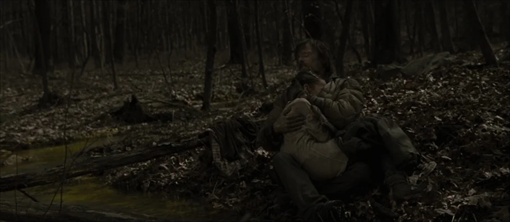 The Road (2009) movie still: medium — A dark, leafless forest floor with a small muddy stream running alongside the ground. A man sits by the water…