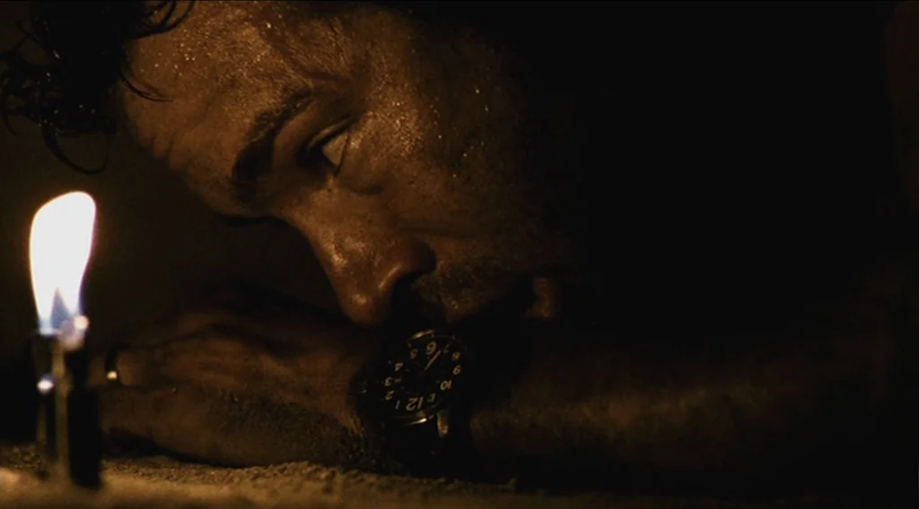 Buried (2010) movie still: closeup — A dark, nocturnal outdoor environment with sand visible in the foreground. A man lies prone while looking at a…