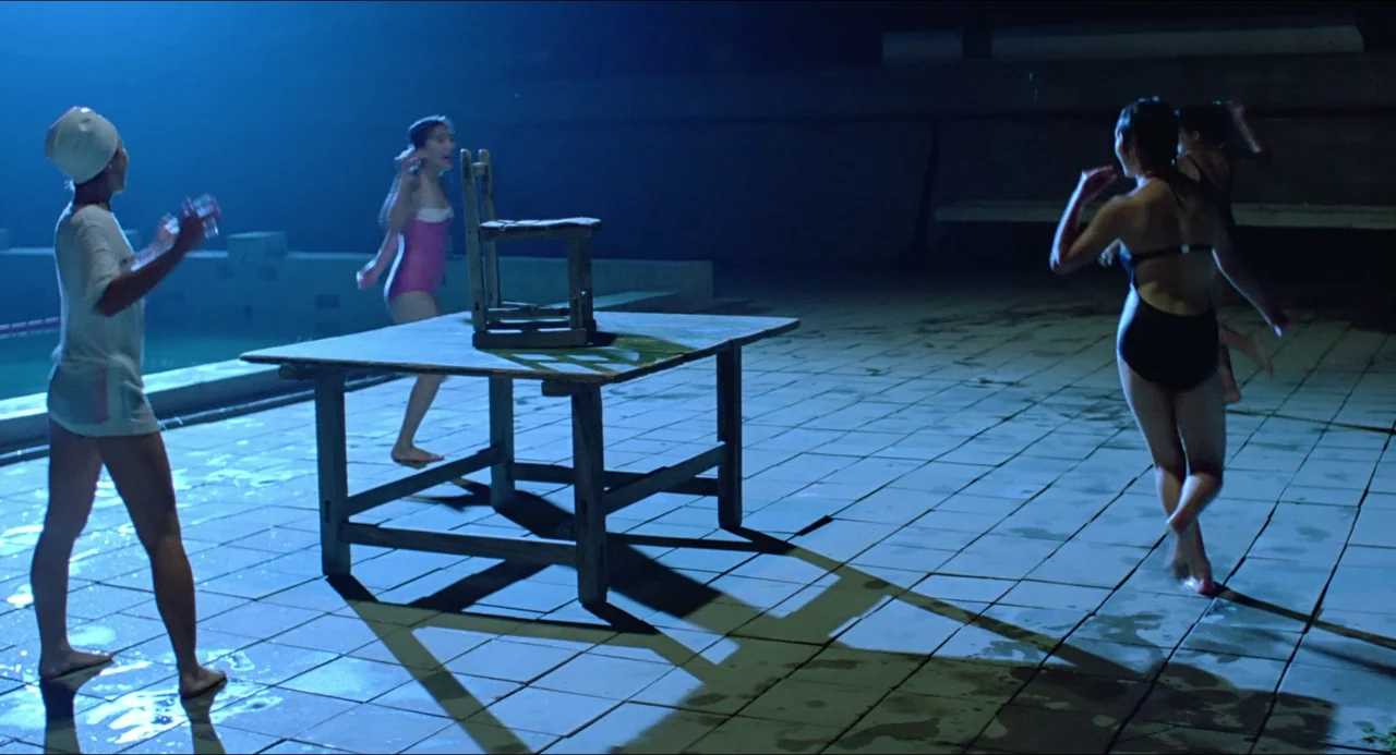 Typhoon Club (1985) movie still: wide — An outdoor pool deck at night features a wooden table holding a single chair. Three women move around the…