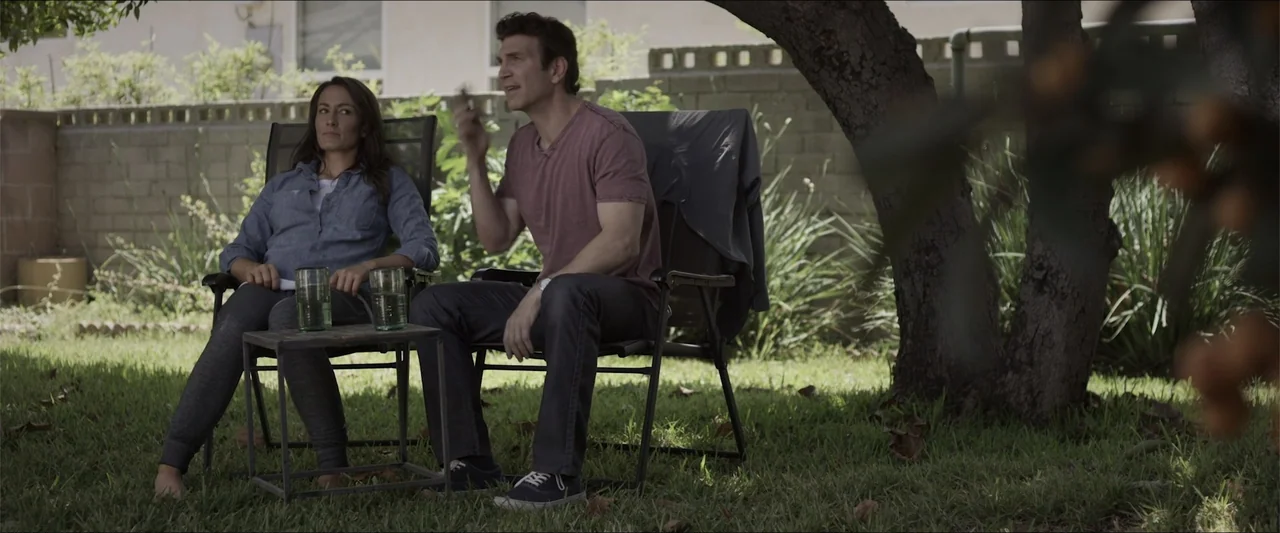 Heavenly Deposit (2019) movie still: medium — A residential backyard with green grass and a large tree canopy overhead. A man and a woman sit in…