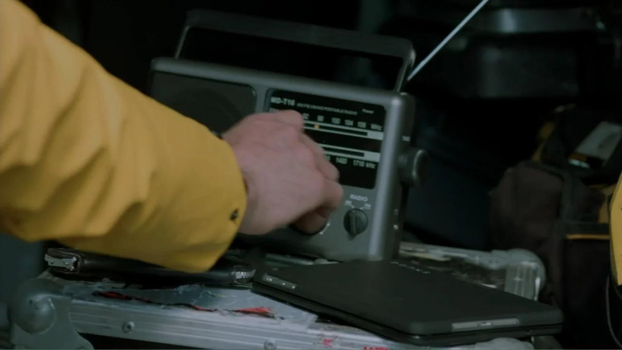 Storm Damage (2000) movie still: closeup — A dark indoor space features a grey portable radio resting on a metal equipment case. A hand wearing a…