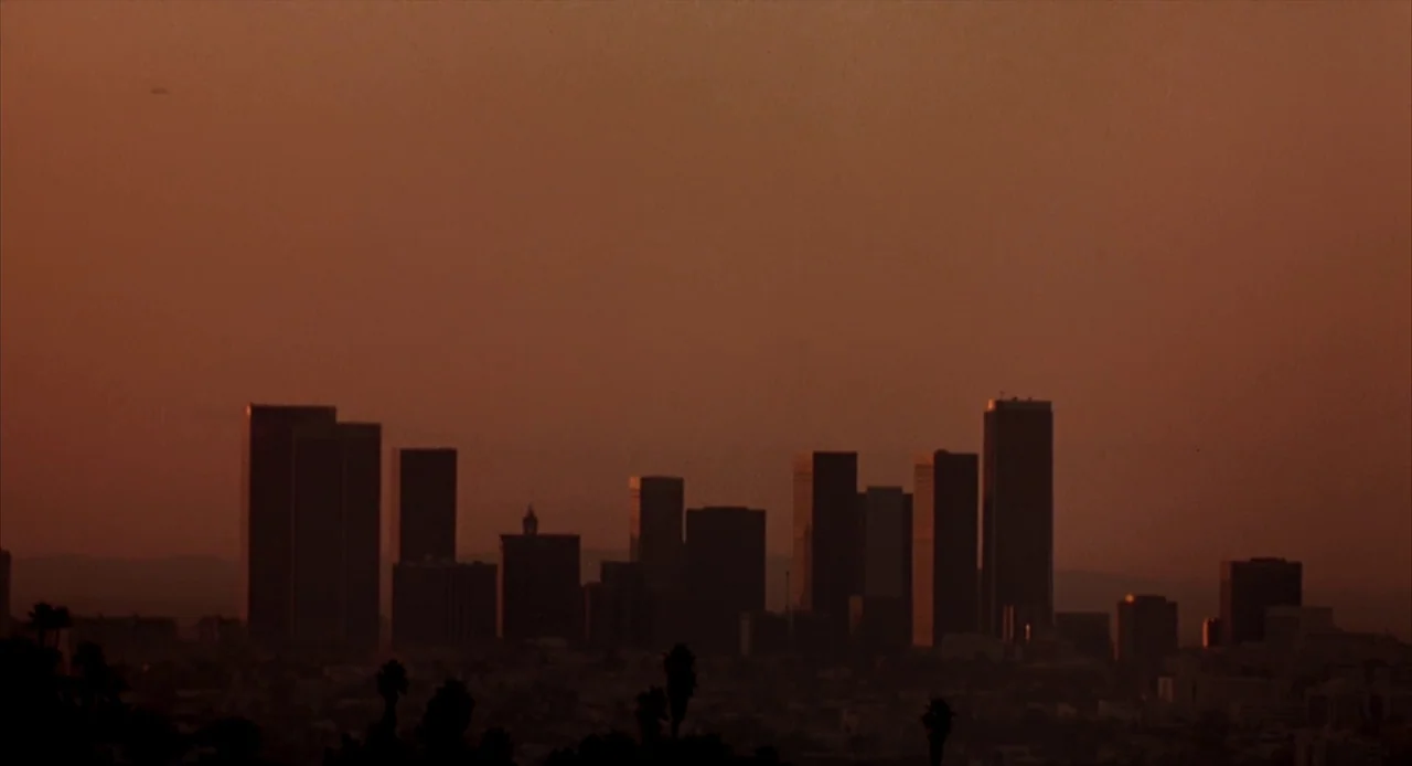 To Live and Die in L.A. (1985) movie still: establishing — The Los Angeles skyline is seen silhouetted against a dark, hazy orange sunset. Low-lying…
