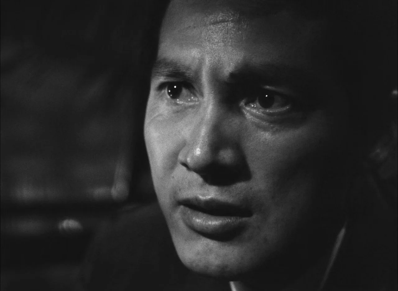 Hiroshima Mon Amour (1959) movie still: closeup — A man stands in a dimly lit, nondescript interior space with high-contrast lighting. The man is…