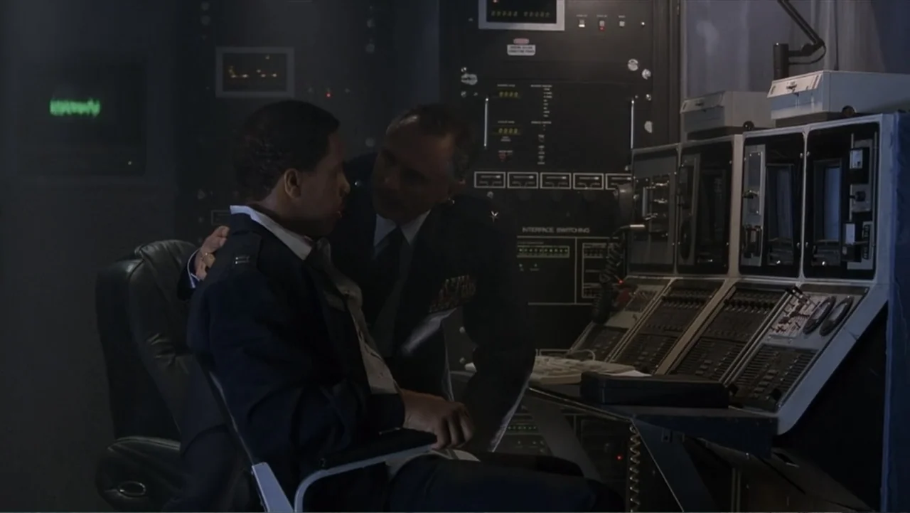 Crisis Point (2012) movie still: medium — A dark military control room contains large server racks and glowing computer console monitors. An officer…