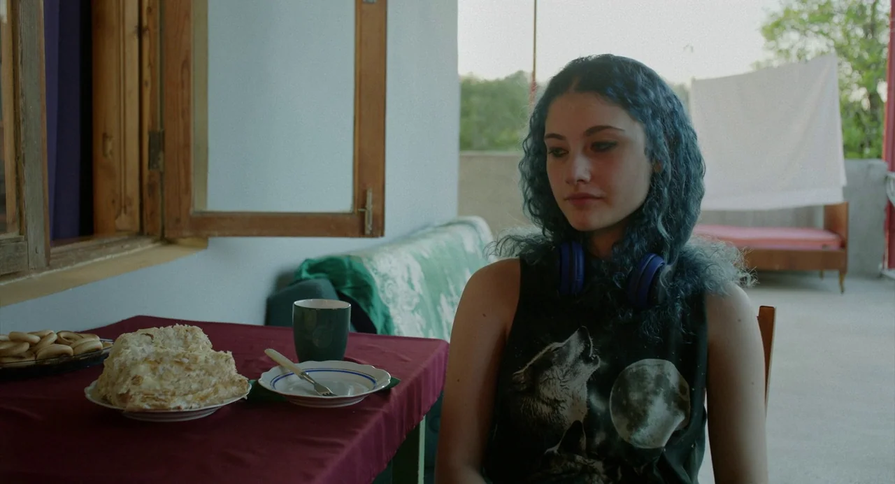 Blackbird Blackbird Blackberry (2023) movie still: medium — A teenage girl sits on a covered porch area during the daytime. A wooden table holds a…