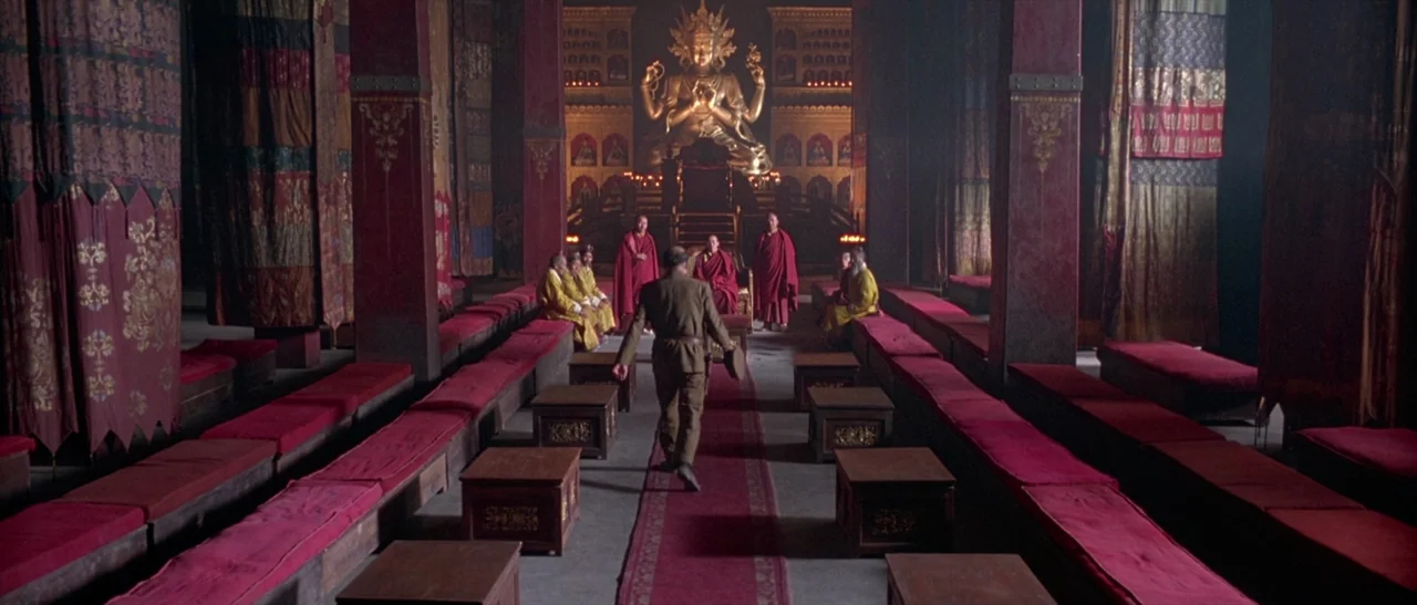 Kundun (1997) movie still: wide — A spacious temple interior with red seating rows and hanging decorative textile tapestries. A man in uniform walks…