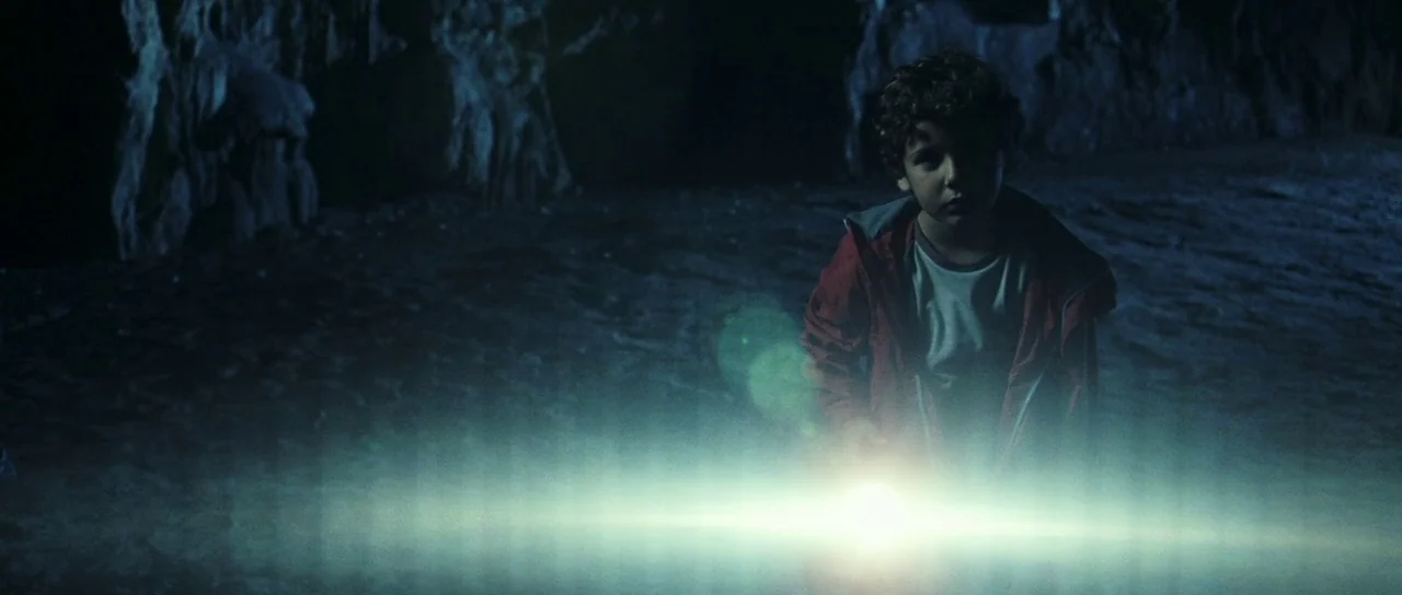 The Orphanage (2007) movie still: medium — A dark, rocky underground cave interior with stalactite formations visible in the background. A young boy…