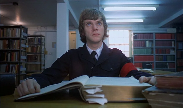 A Clockwork Orange (1971) movie still: medium — A quiet institutional space filled with rows of metal shelves packed with books. A young man wearing…
