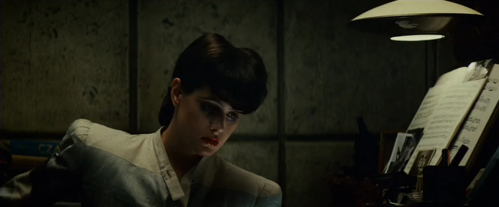 Blade Runner (1982) movie still: medium — A dimly lit room features a woman seated beside a desk with music sheets. She looks downward while…