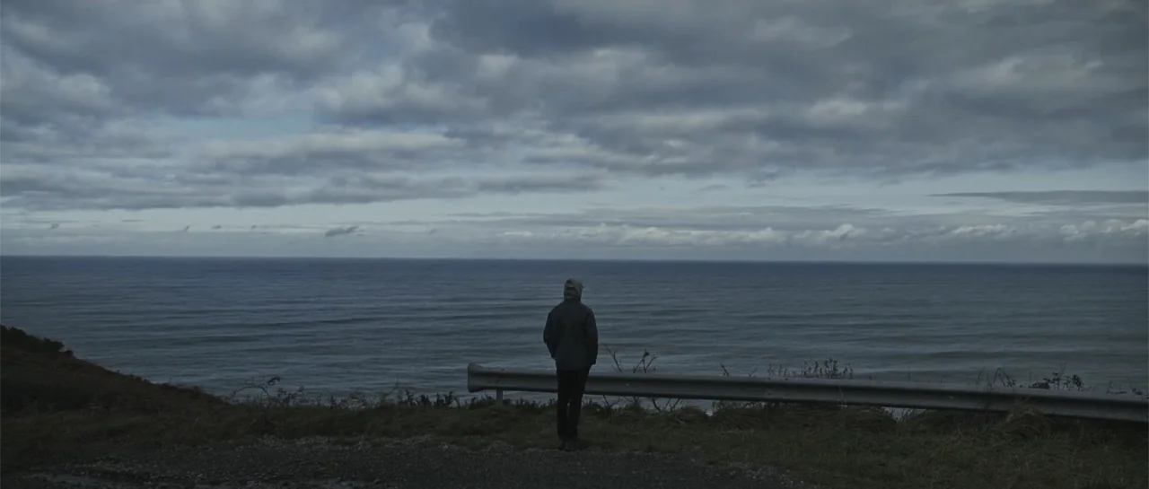 Taking Back the Waves (2017) movie still: wide — A grassy clifftop overlooks a vast ocean under a heavy, overcast grey sky. A lone person in dark…
