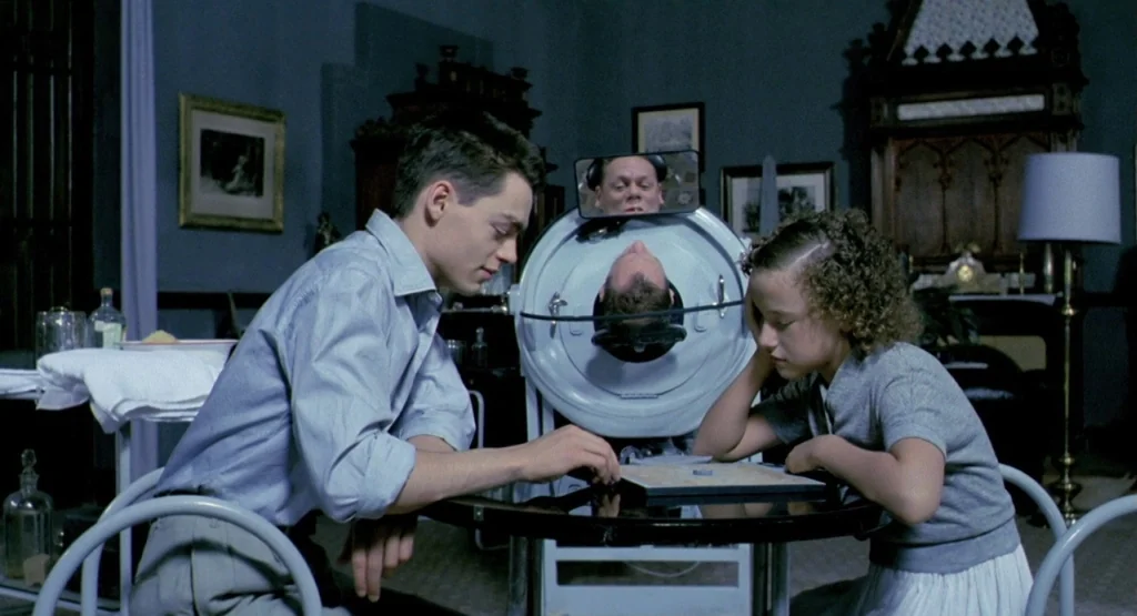 In a Glass Cage (1986) movie still: medium — A dimly lit, vintage hospital room contains an iron lung device in the background. A young man and a…