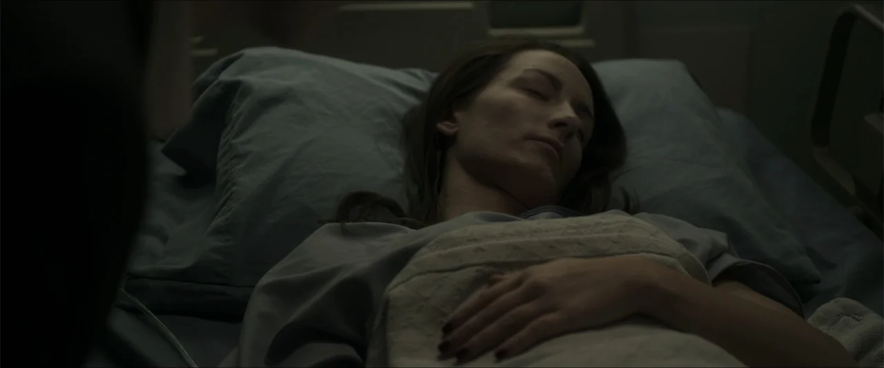 Heavenly Deposit (2019) movie still: medium — A dimly lit hospital room with a patient bed and blue pillows. A woman with long dark hair rests in…