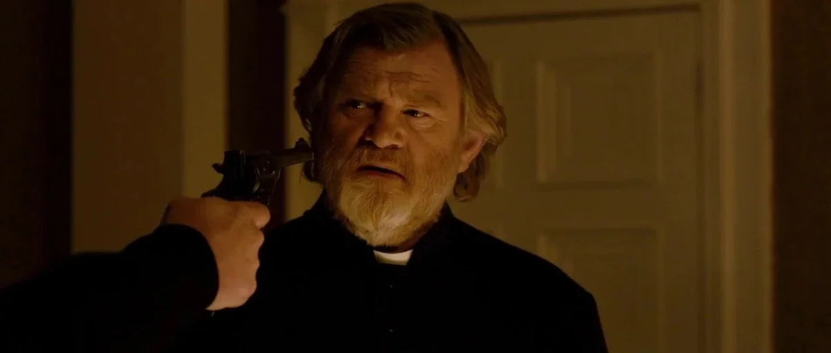 Calvary (2014) movie still: medium — A dimly lit room features a doorway in the blurred background behind the subject. A person points a black…