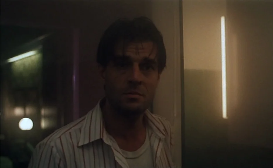 The Fourth Man (1983) movie still: medium — A dimly lit interior hallway with a vertical light fixture glowing on the wall. A man with tired eyes…