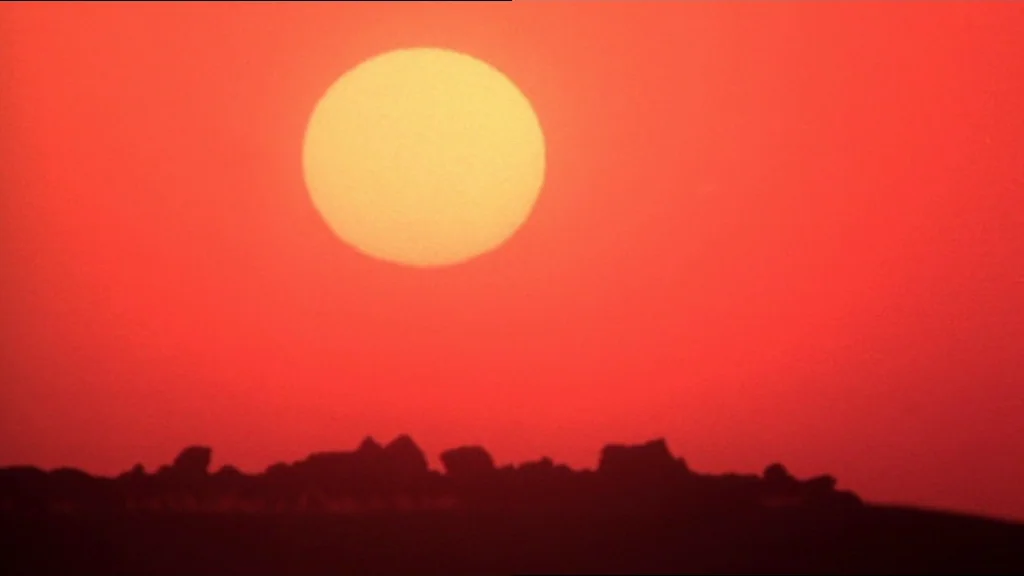 The Exorcist (1973) movie still: establishing — A vast, barren landscape features a rocky horizon silhouette against a vivid sunset sky. The bright…