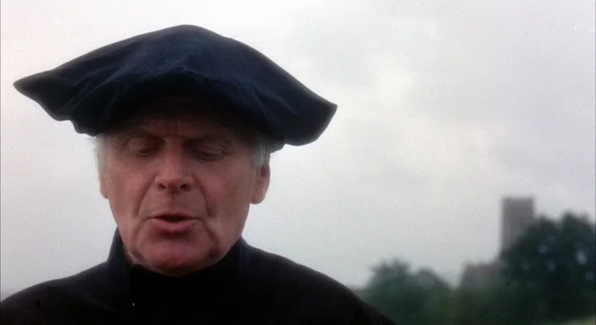 Witchfinder General (1968) movie still: closeup — A man wearing a black cap stands in an open field under a cloudy sky. In the blurry background, a…