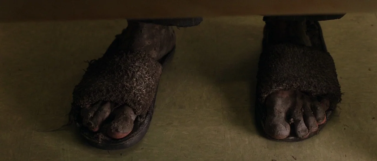 Halloween (2007) movie still: closeup — A pair of mud-caked feet rest on a tiled floor inside a small enclosure. The feet are inside dark, worn-out…